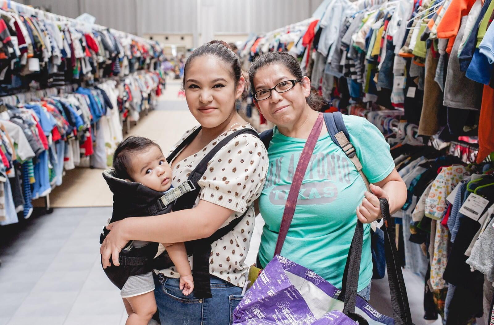 Three generations shop together at their local JBF sale. Mom carries baby in a baby carrier while grandmom stands behind them.