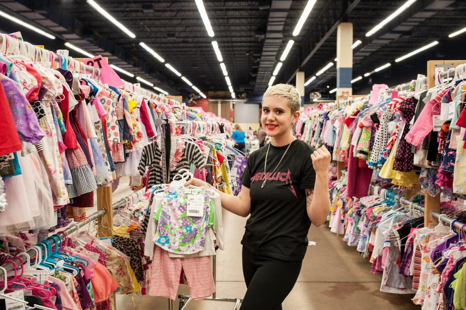 A young JBF shopper mom in a black Metallica T-shirt holds an outfit she intends to buy at her local JBF sale.